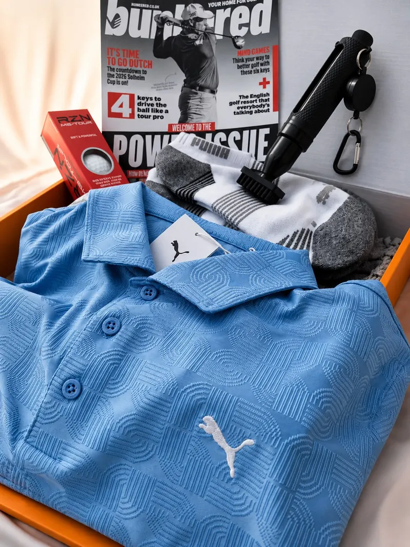 August 2026 box seen from above and to one side: a light blue textured Puma polo with its hangtag tucked into an open orange Airdropgolf box, with a red sleeve of RZN MS-Tour golf balls, a copy of bunkered golf magazine, white and grey Puma socks and a black club-cleaning brush.