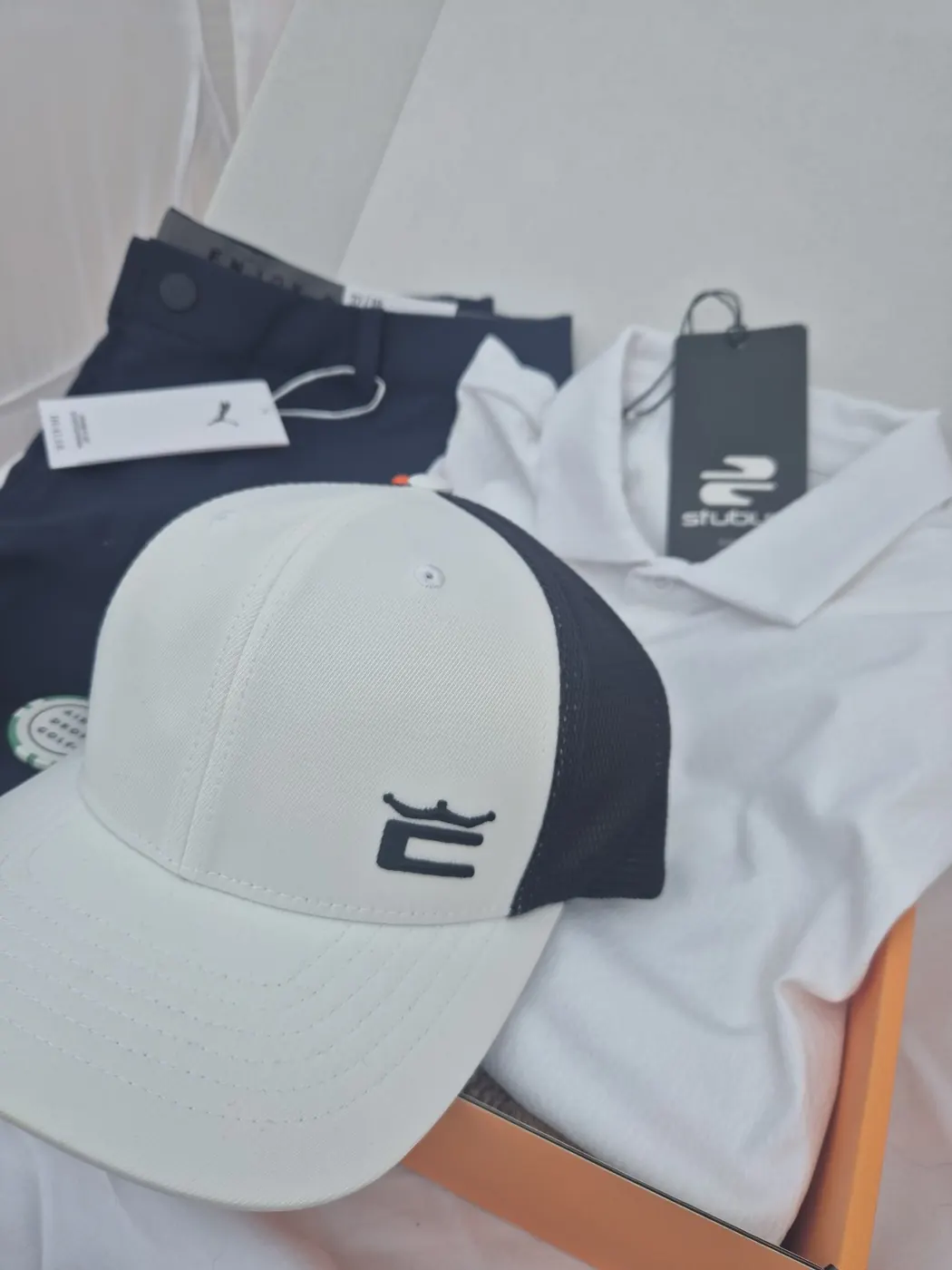 July 2026 box from a second angle: a white and navy mesh-back Cobra cap in front of a white Stuburt polo with hangtag and navy Puma trousers with a white Puma tag, with a green Airdropgolf poker-chip ball marker, in the orange Airdropgolf box.