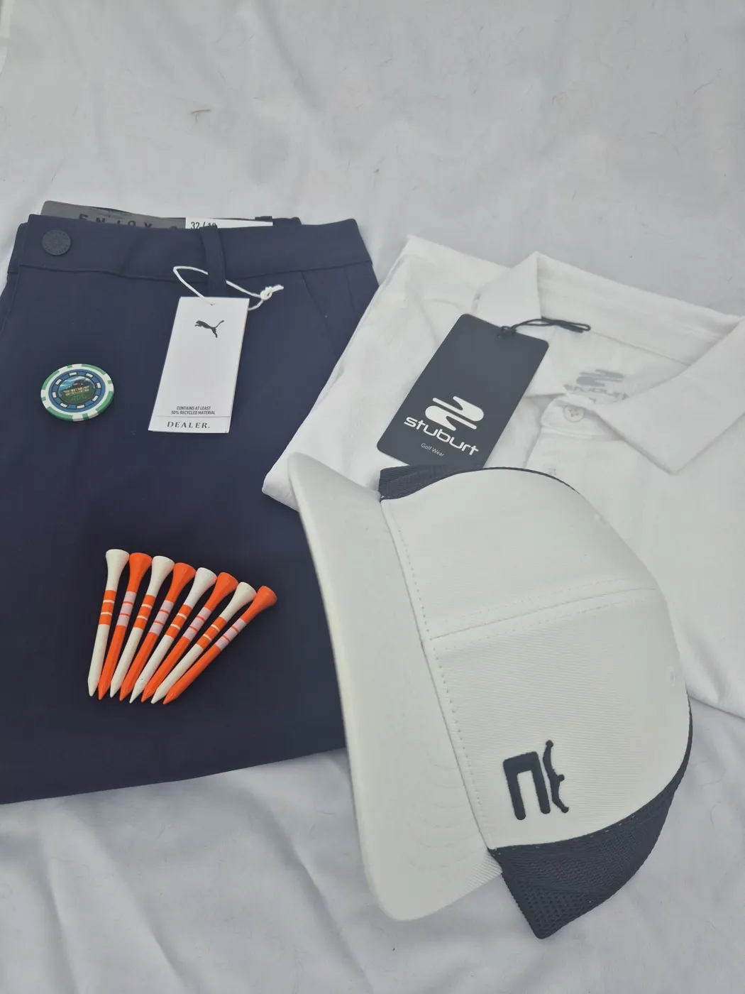 July 2026 box contents laid on a white cloth: folded navy Puma Dealer trousers with hangtag, a white Stuburt polo with its black Stuburt tag, a white and navy mesh-back cap, a green poker-chip ball marker and a fan of orange and white tees.
