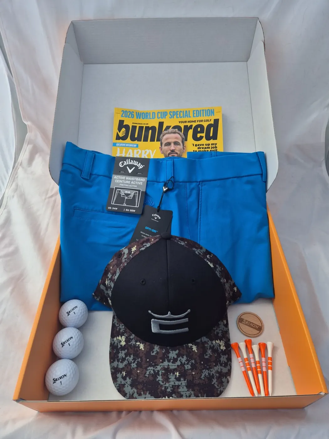 May 2026 box laid in the open orange Airdropgolf box: bright blue Callaway trousers with an Opti-Dri hangtag, a black and camo Cobra cap, three Srixon golf balls, a wooden Srixon ball marker, orange and white tees and a bunkered 2026 World Cup special edition magazine.